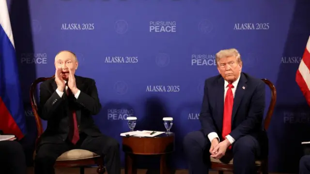 Putin and Trump watch next to each other inside, in front of a backdrop that says "Pursuing Peace" and "Alaska 2025".